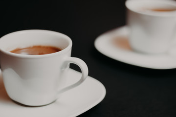 White color cup of coffee with saucer on black background, another one in blurred background, focus on the handle of first cup