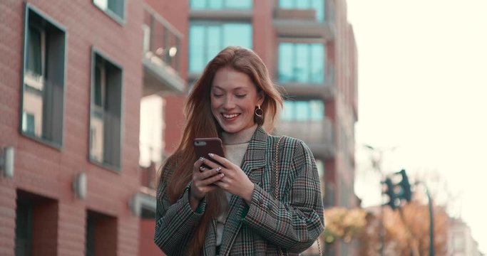 Attractive Girl having a Walk in the Town while Typing Messages on her Smartphone. Having online Conversation. Looking at screen of her Mobile. Woman with beautiful hair and charming look