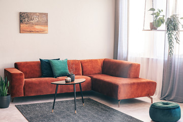 Real photo of a red, suede corner couch standing against white wall with an oil painting on it in living room interior
