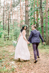 Back view of stylish rustic couple of newlyweds on their wedding day, walking and running in the forest. Happy young bride, elegant groom holding hands and having fun