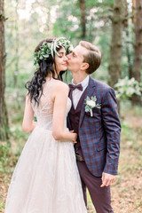 Bride in wreath on head and elegant dress and groom in stylish suit walking in forest kissing. Happy newlyweds at a wedding in nature, green pine forest. Rustic Wedding Couple