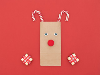 Christmas deer made of craft shopping bag and two Xmas canes with gift boxes on red background. New Year greeting card. Christmas and New Year concept. Flat lay style with copy space.