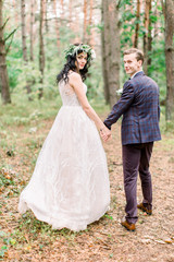 Back view of happy smiling stylish bride in floral wreath and handsome groom walking in the forest, having fun on the their wedding day
