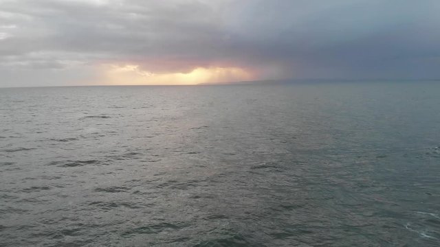 An aerial backward footage of crashing waves on a choppy sea under a stormy grey sky with sun glowing behind clouds