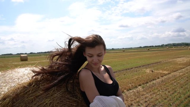Attractive sensual brunette girl in black undershirt stands in the sun near the round haystack among the field, hair fluttering in the wind, blue sky with clouds on the background