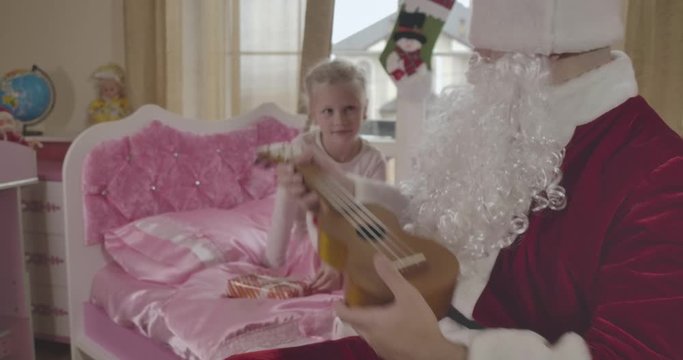 Cute Caucasian child sitting on pink bed and looking at Santa playing ukulele. Adult man in Santa Claus costume giving her musical instrument. Holidays, miracle, happiness. Cinema 4k ProRes HQ.