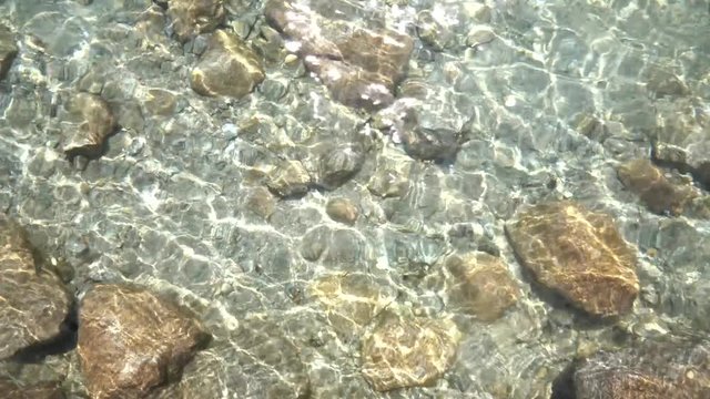 Big stones under clear transparent water with waves and glare of the sun