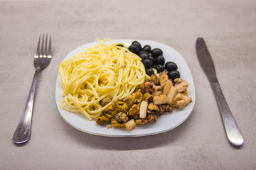 white plate on a gray kitchen table with spaghetti with black olives and seafood knife and fork on the sides