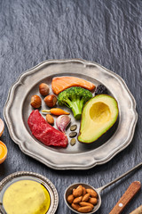 Plate with a ketogenic diet food. Beef, salmon, egg, broccoli, tomato, nuts, carrots, mushrooms, cucumber, dates. Diet food on a dark background with copy space. 
