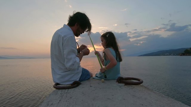 Rear view of little girl sitting on the edge of a dock and fishing with father Mediterranean Sea at sunset. Enjoying adventure in summer vacation