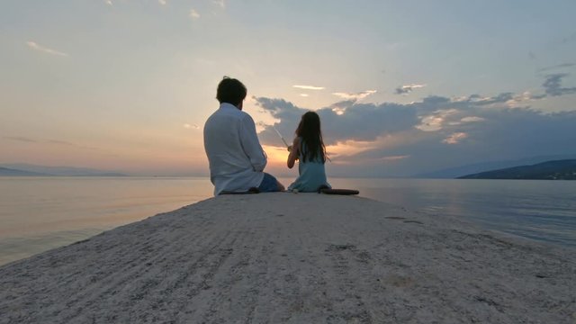 Rear view of little girl sitting on the edge of a dock and fishing with father Mediterranean Sea at sunset. Enjoying adventure in summer vacation