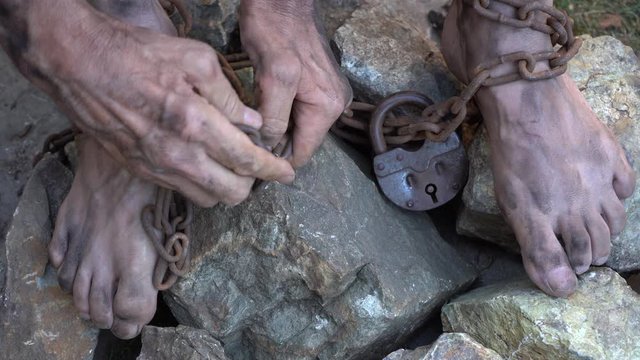 Man in captivity. A slave in iron chains is trying to free himself. A scene from the life of a slave. The symbol of slave labor. Hands and feet in chains. Breaking chains