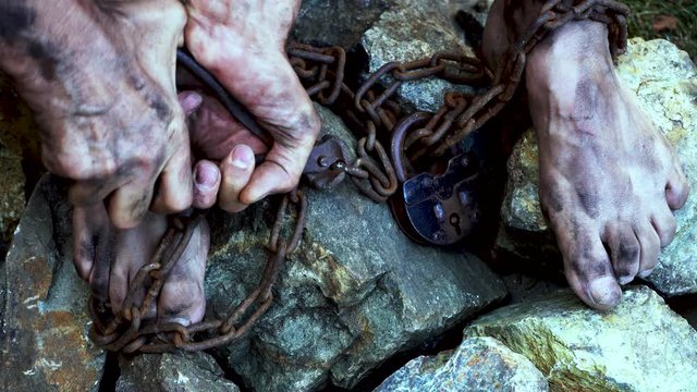 Man in captivity. A slave in iron chains is trying to free himself. A scene from the life of a slave. The symbol of slave labor. Hands and feet in chains. Breaking chains