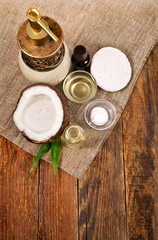 Half coconut with bottle and cup of coconut oil, cosmetic dispenser, sponge and cosmetic cream on wooden background. Top view. Text space.
