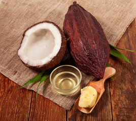 Half coconut with bottle of coconut oil, cocoa butter in a wooden scoop and cocoa pod with palm leaves on a wooden background.