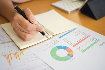 Close shot of businesswoman hands holding a pen writing something on the paper on the foreground in office. Recording concept. Using smartphone tablet. Freelance Work, Business concept