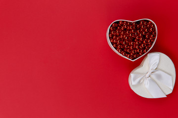 FLAT LAY STYLE. On a red paper surface on the right is a white heart-shaped box with red beads. There is a lot of free space, horizontal, close-up. Valentine's Day.