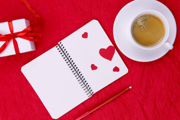 FLAT LAY STYLE. On a red crumpled paper surface there is a white notebook on springs, a red pen, a white box with a gift and coffee in a white cup. Free space, horizontal, close-up. Valentine's Day.