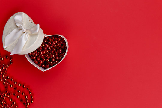 FLAT LAY STYLE. On a red paper surface lies a white heart-shaped box with red beads. There is a lot of free space, horizontal, close-up. Valentine's Day.