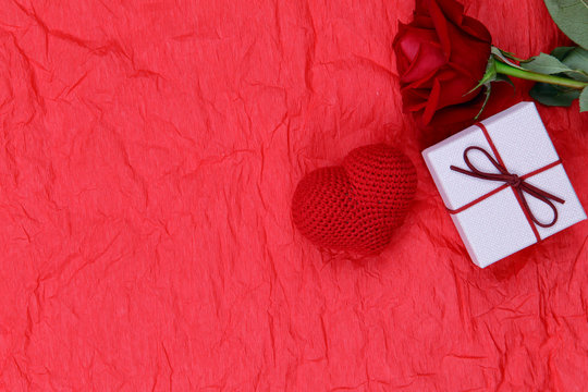 FLAT LAY STYLE. On a red crumpled paper surface there is a white box with a gift, a rose and a knitted heart. A lot of free space, horizontal, close-up. Valentine's day.