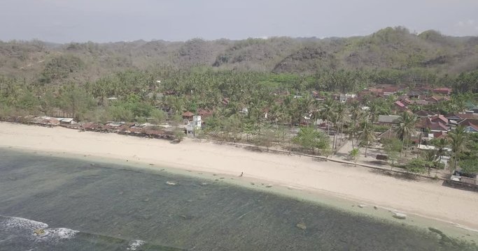 8. Aerial View, Bird View Of Two People Walking On A Beautiful White Sand Beach In Front Of A Village With Many Coconut Trees With A Background Of Hills, Raw Footage, Ungraded, No Color, Log Profile