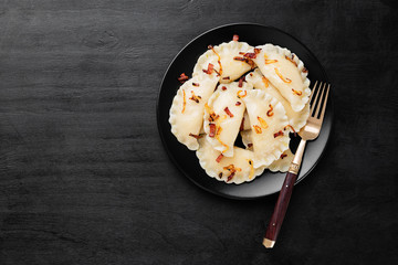 Homemade boiled dumplings filled with potato served with fried onion and bacon on black wooden background.