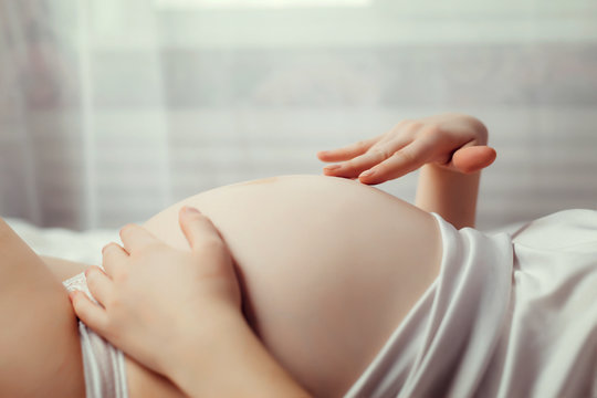 A beautiful pregnant woman in white underwear is lying on her bed and gently stroking her belly. Great time to wait for the baby. Close-up photo of a pregnant belly