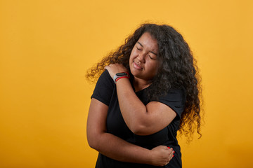 Afro-american young lady with overweight over isolated orange background wearing fashion black shirt keeping hand on shoulder, having pain in muscles. People lifestyle concept.