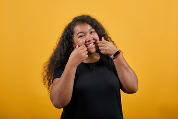 Young woman with overweight pulls smile with fingers, looking at camera over isolated orange background wearing fashion black shirt. People lifestyle concept.