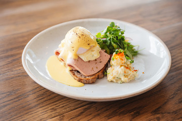 Eggs Benedict with ham, toast and mashed potato. Served with salad on a white plate on wooden table.