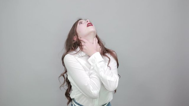 Attractive young woman in fashion white shirt holding hands on neck, choking herself, looking up isolated on gray background in studio. People sincere emotions, lifestyle concept.
