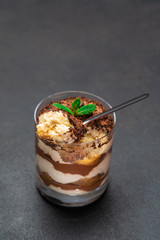 Classic tiramisu dessert in a glass cup and pieces of chocolate on dark concrete background