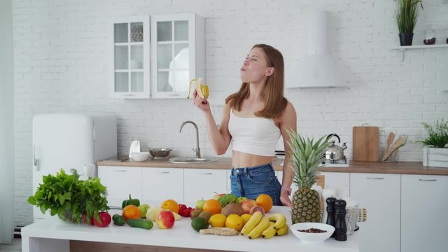 Girl Eating Banana Indoors. Young Woman On The Modern Kitchen Background. Healthy Fruit And Vegetables On The Table. Attractive Female Vegetarian Is Eating Fruit.