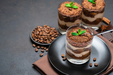 Classic tiramisu dessert in a glass cup and pieces of chocolate on dark concrete background