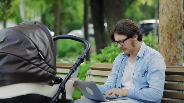 Young man swinging baby stroller, working on laptop in city park, freelancer