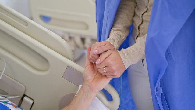 Close-up woman's hands supporting sick patient. Female visitor holds hands of her father in the hospital room. Senior male patient is laying in bed in clinic.