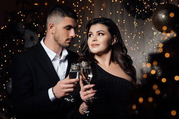 Loving couple, is stylishly dressed near a Christmas tree. Christmas time. Christmas mood. Loving couple celebrating New Year and Christmas with champagne. A bearded guy with a girl pose in a studio 