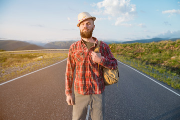 Portrait of a bearded happy smiling traveler hipster with a backpack in a plaid shirt and a hat next to an unknown car stands on the road at sunset in the mountains. Happy and confident travel concept