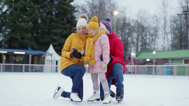 Zoom in of happy family of mother, father and little daughter posing for selfie on smartphone at outdoor ice skating rink on cold winter day