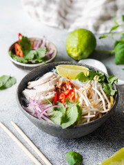 Traditional Vietnamese soup- pho ga in bowl with chicken and rice noodles, mint and cilantro, red onion, chili, bean sprouts and lime on grey background. Asian food. Copy space.