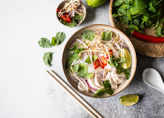 Traditional Vietnamese soup- pho ga in bowl with chicken and rice noodles, mint and cilantro, red onion, chili, bean sprouts and lime on grey background. Asian food. Copy space. Top view - obrazy, fototapety, plakaty
