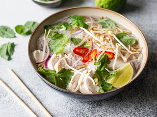 Traditional Vietnamese soup- pho ga in bowl with chicken and rice noodles, mint and cilantro, red onion, chili, bean sprouts and lime on grey background. Asian food.