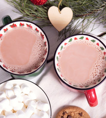 two mugs red and green with cocoa and marshmallows, homemade cookies on a napkin, spruce branches on a light background, top view