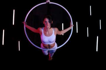 Young woman performing acrobatic element on aerial ring against dark background