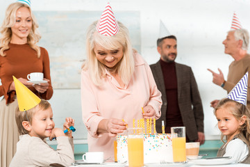 happy senior woman putting candles in birthday cake near family in party caps