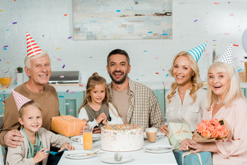 happy family sitting at kitchen table near birthday cake under falling confetti