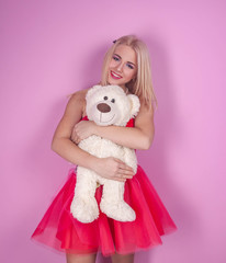 beautiful happy blonde in a bright pink dress looks at the camera, hugs a white toy bear, on a pink background