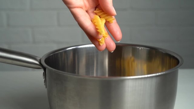 Hand throwing fusilli pasta into metal pot, white brick wall background. Culinary, cooking, recipe concept. Slow motion, close-up