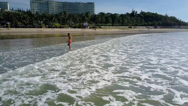 Amazing view rolling ocean wave follows girl in red swimsuit going to sand beach against tropical palm park and hotels