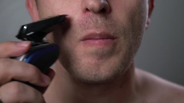 A man shaves his beard and mustache with an electric razor at home. Hair removal in front of a mirror. Men's care. Brutal man with a big beard. Change of image. Dry shave. Electric shaver.